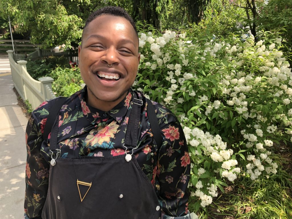 Photo of a smiling Black person wearing long-sleeved floral and black overalls. They have a septum ring and stand in front of a blooming flower bush with white buds and green leaves.